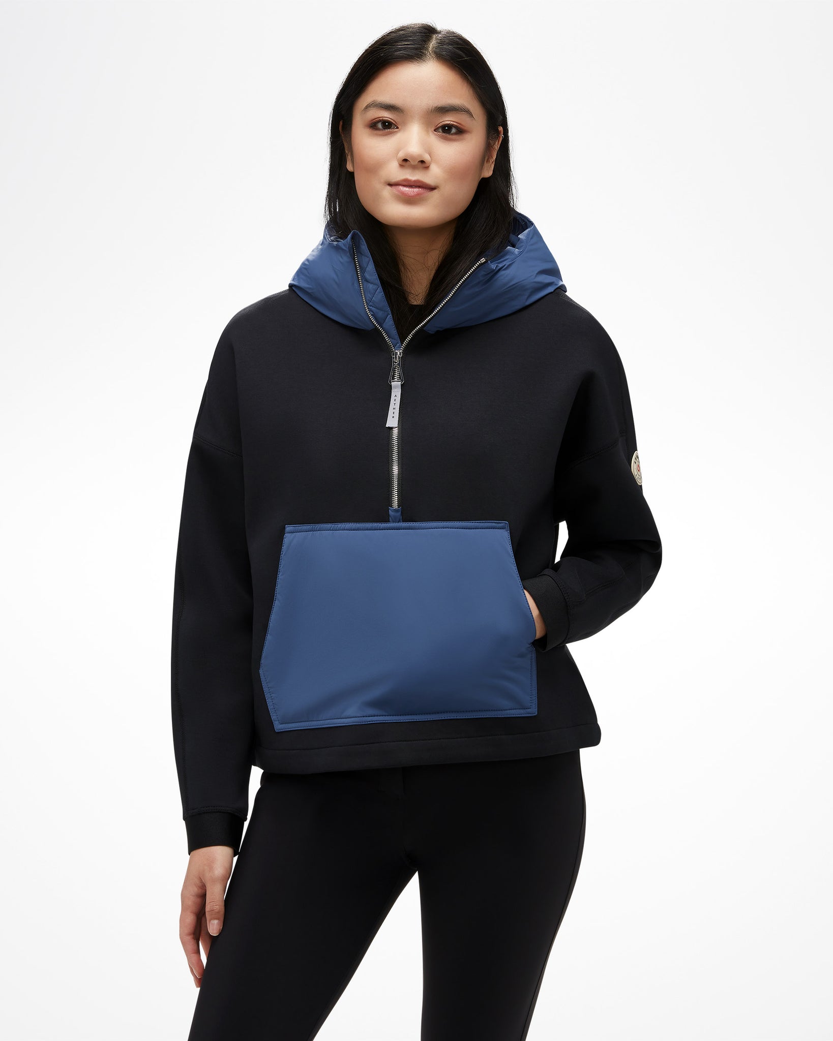 Aspen Collection Aether Align Hooded Anorak, half zip. Black base with blue puffy kangaroo sweater, blue puffy hood, and silver zipper detail, view from front on model.