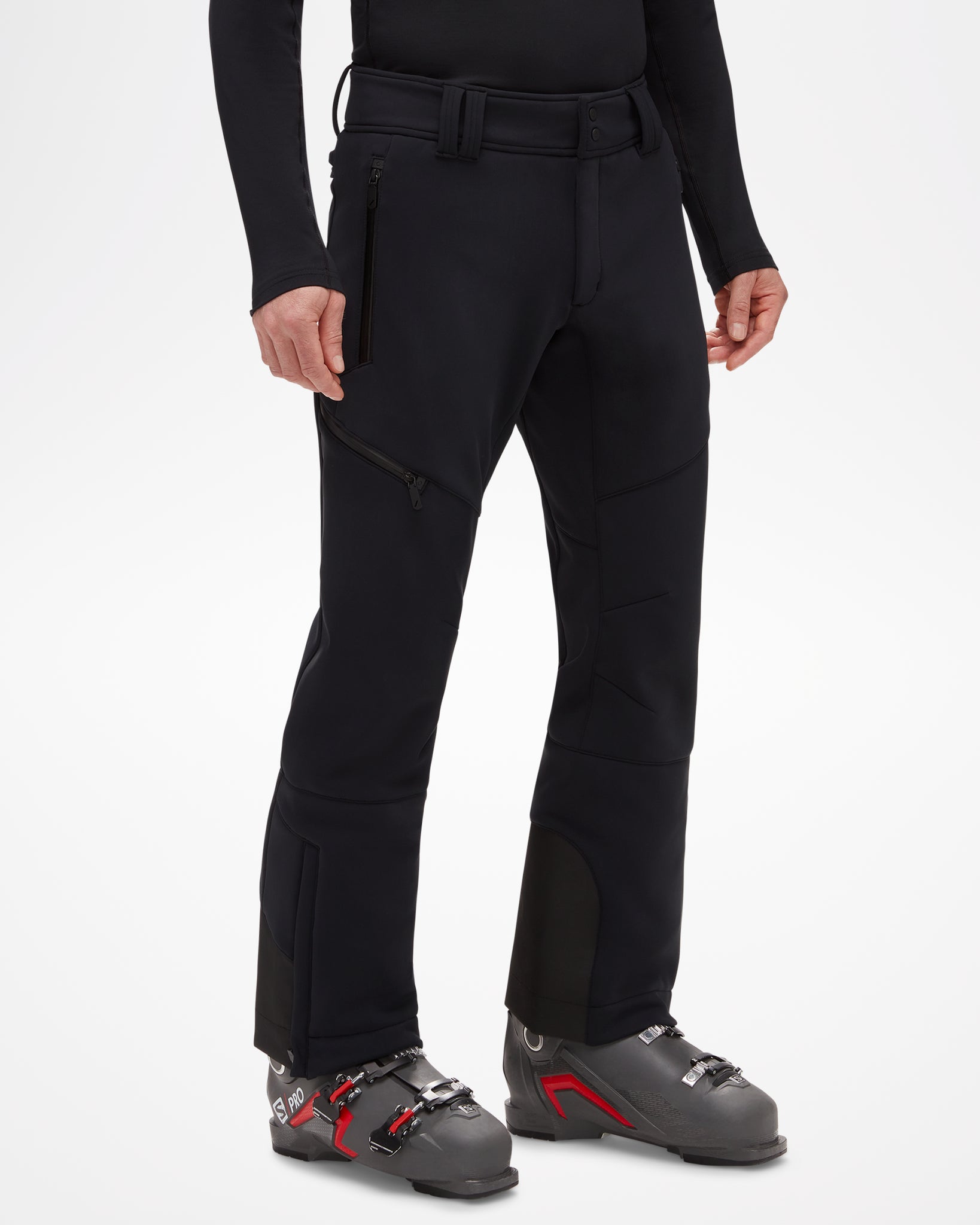 Beattie Softshell Men's Ski Pant, modeled with ski boots, all black. Slightly fitted around waist and thigh, fanning out slightly below knee, front-side view