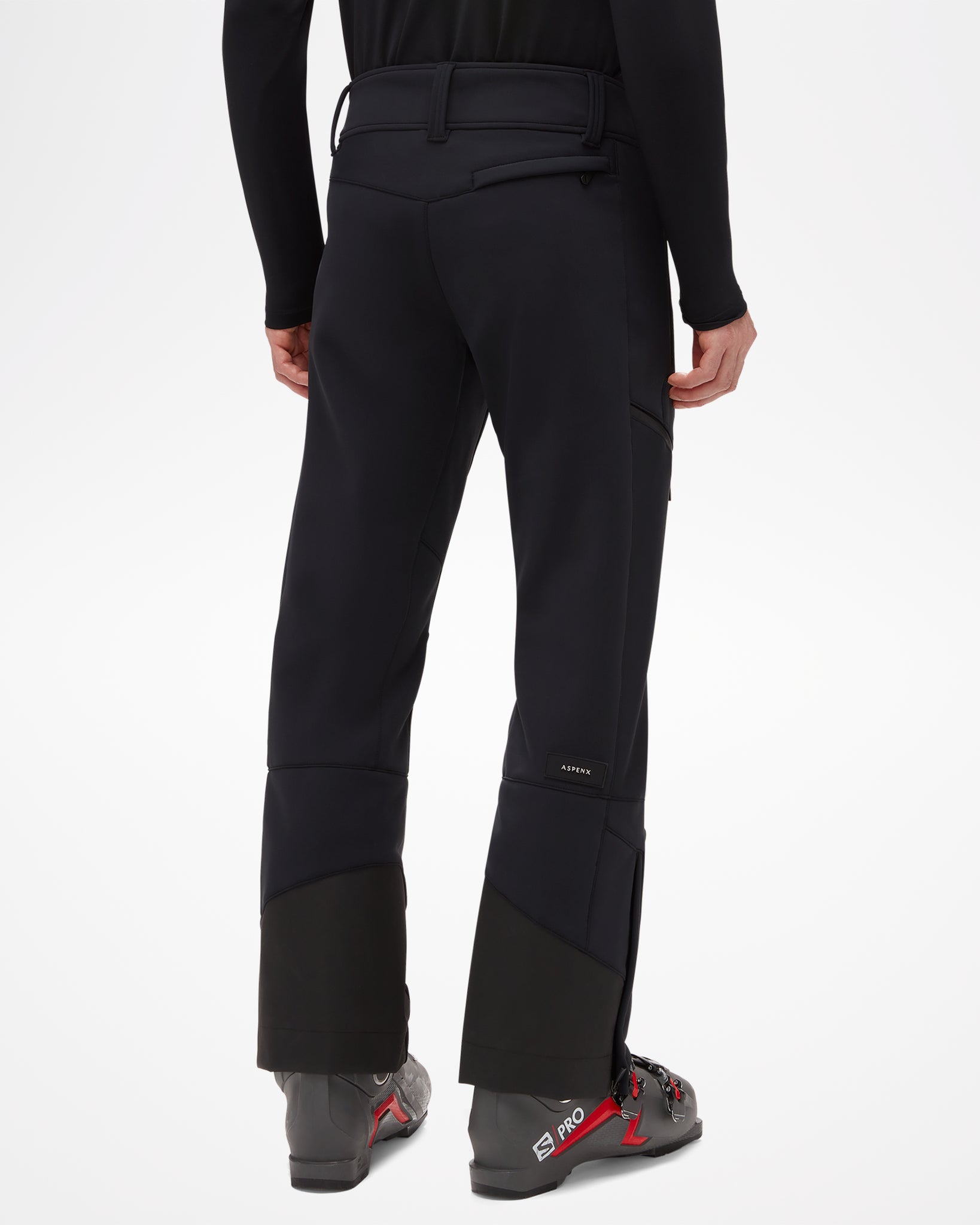 Beattie Softshell Men's Ski Pant, modeled with ski boots, all black. Slightly fitted around waist and thigh, fanning out slightly below knee, back view of pants