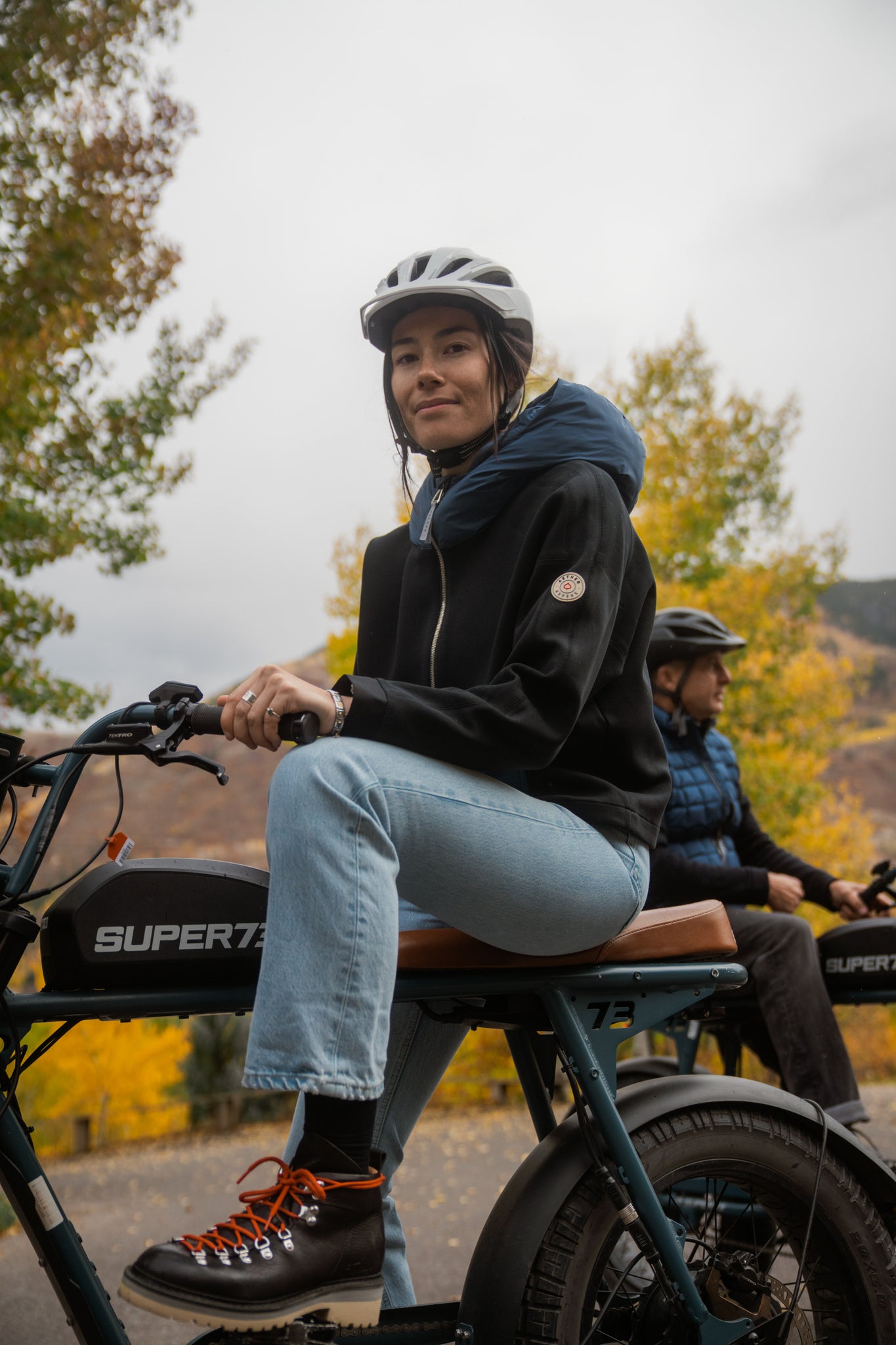 Aspen Collection Aether Align Hooded Anorak, on model with blue jeans. She is on an electric bike, wearing the black and dark blue hoodie on a cloudy fall day.